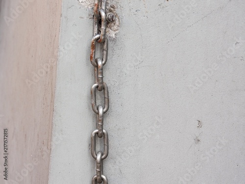 Vertical steel metal link chain anchored to a concrete wall surface with slight rust spots.
