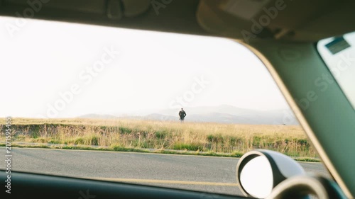 Person standing outside a car on a wide field during daytime with a clear sky and distant mountains