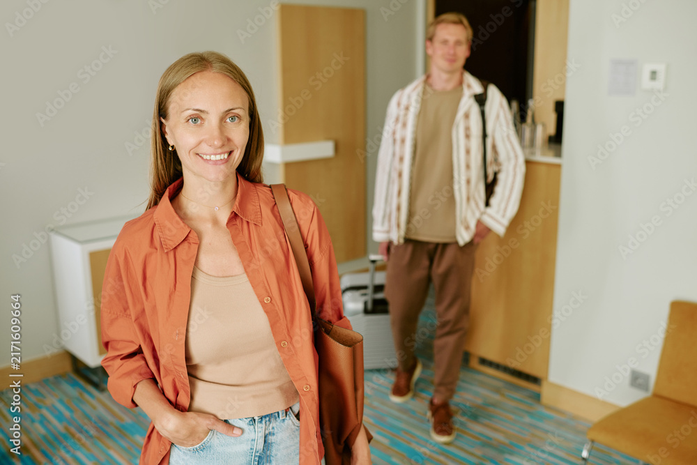 Fototapeta premium Portrait of smiling Caucasian young adult woman standing in hotel room with man in background. Image represents travel and hospitality, ideal for tourism marketing campaigns