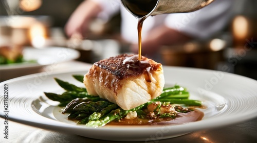 Gourmet seared white fish fillet served on fresh green asparagus with rich brown sauce being poured in professional restaurant kitchen setting