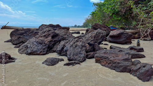 Quadro su tela Ground-level photo showing a tight group of heavy weathered rocks on a sandy beach beside a dense tropical forest background