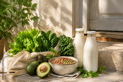 Doorstep with avocado, leafy greens, legumes, plant-based milk bottles, vegan grocery concept