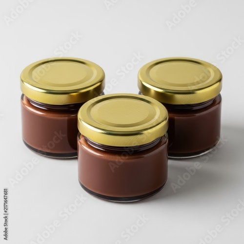 Three small clear glass containers containing smooth dark chocolate cream, sealed with reflective gold metal caps on a neutral surface, spreadable, reflective, delicious