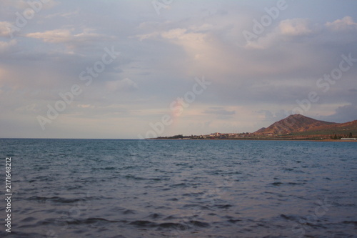 Canvastavla Tranquil lake landscape with gentle waves under a cloudy sky and a faint rainbow over distant mountainous coastline during dusk