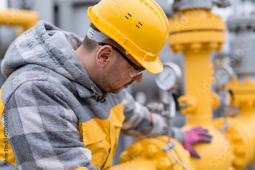 Worker in hard hat inspects yellow gas pipeline equipment. Focused, he checks valves with gloved hands. Industrial setting suggests maintenance or safety inspection