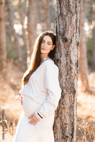 Stylish pregnant woman 30-35year old wearing knitted maternity clothes posing with closed eyes outdoors over autumn nature background. Motherhood.