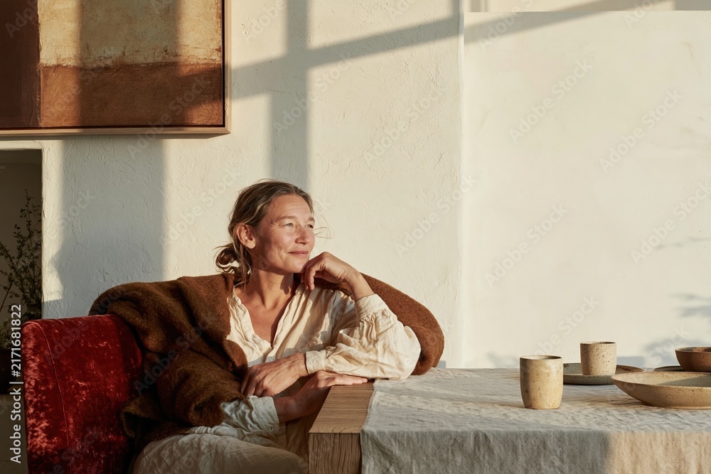 Fototapeta premium Mature woman resting in a cozy dining room, wearing a warm wool blanket with a thoughtful expression, featuring soft light and copy space