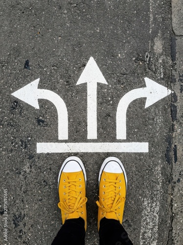 Person in Yellow Sneakers Standing Before Three Directional Arrows on Asphalt Representing Choice and Decision Making
