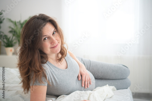 Pregnant woman reclining on bed in casual maternity lounging. Expecting mother gazing at camera with serene expression and crossed legs. Future parent enjoying peaceful soft moment in bright bedroom.