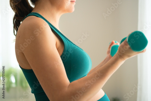Pregnant woman stands holding dumbbell, exercising with strong body empowerment by window. Athlete and mother shows confident, fit determination during indoor strength training in home gym