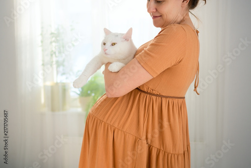 A pregnant woman holds a white cat in a bright room. The expectant mother hugging her companion shows compatible, careful handling and compatibility with pets during pregnancy at home.
