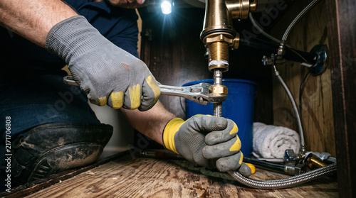 Professional plumber fixing kitchen sink piping using metal wrench under dark cabinet with bright task lighting