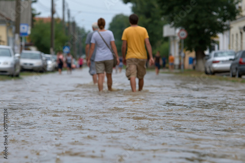 Two people wade through shallow floodwaters. They walk side by side  facing away from the camera. Streetcars and parked cars line the submerged road. Trees and buildings frame the background