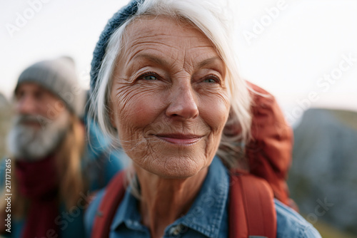 Sunlit trail. Senior woman smiles warmly. Her white hair glows gently. Soft background hints at nature. She carries a red backpack. Adventure and peace meet here