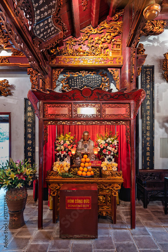 Ornate altar with a statue, flower arrangements, and orange offerings inside the historic Quan Thanh Temple in Hanoi, Vietnam, featuring a wooden donation box in the foreground