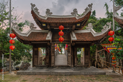 Traditional wooden entrance gate of Thuy Trung Tien Temple decorated with red lanterns and a stone bridge leading to the pagoda in Hanoi, Vietnam