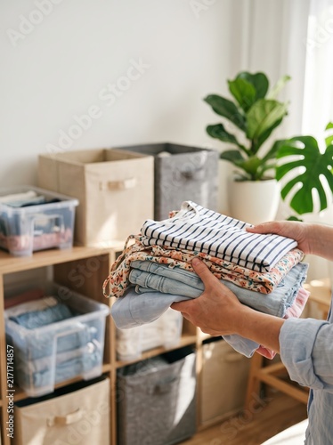 Woman holding stack of folded summer clothes. Clean wardrobe organization and seasonal clothing storage. Household chores, tidying up closet, decluttering and home organizing concept.