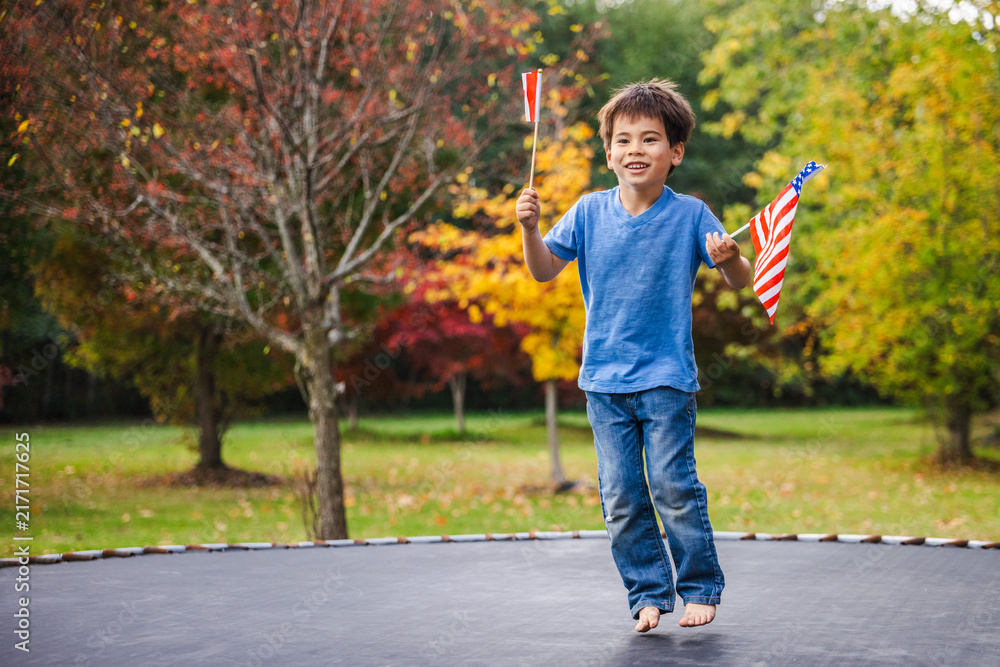 Fototapeta premium Smiling Boy Jumping Barefoot With American and Red-and-White Flags