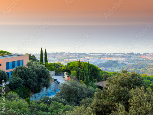 Scenic elevated panoramic view over Mataro coastal landscape at sunset with modern villa nestled in lush green Mediterranean trees and hazy calm sea horizon.