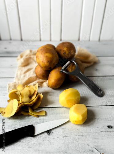 Fotografía Peeling raw potatoes with a knife and vegetable peeler on a white wooden table