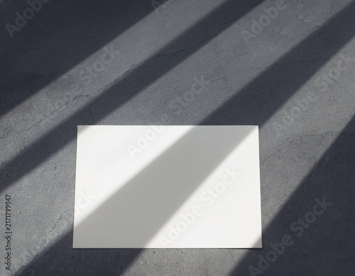 Pristine white blank rectangular paper card rests on a rough dark gray concrete surface illuminated by strong diagonal sunlight shadows creating a minimalist aesthetic background