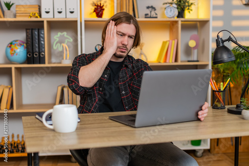 Bild auf Leinwand Young man on home table sees bad laptop result and hits desk in stress then shuts screen with deep sigh