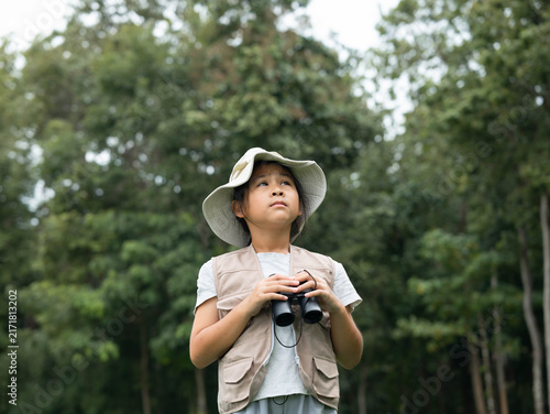 Cute Asian girl in safari hat holding binoculars looking up at treetop in park