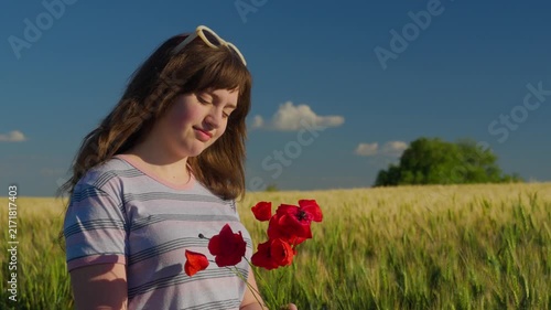A beautiful young teenager smiling peacefully while holding vibrant red poppy flowers in a vast wheat field under a clear blue sky at sunset