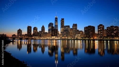 Panoramic view of a city skyline at dusk, with buildings illuminated by lights and reflected in the calm water below.