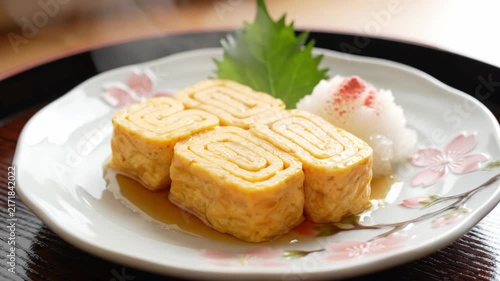 Close-up of freshly prepared tamagoyaki, a Japanese rolled omelet, presented with grated daikon radish and a perilla leaf on a decorative plate with cherry blossom motifs.
