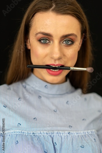 Plakat w ramie Playful young woman holding a makeup brush in her teeth