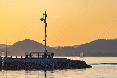Fonyod, Hungary: Harbor pier with a communication tower and a sailboat on Lake Balaton at sunset, with distant hills and Szigliget Castle silhouette.