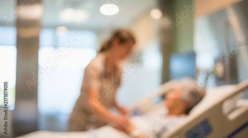 Healthcare professional provides care to a patient resting in a hospital bed. Soft focus background captures a clinical environment with warm ambient lighting and blurred silhouettes.