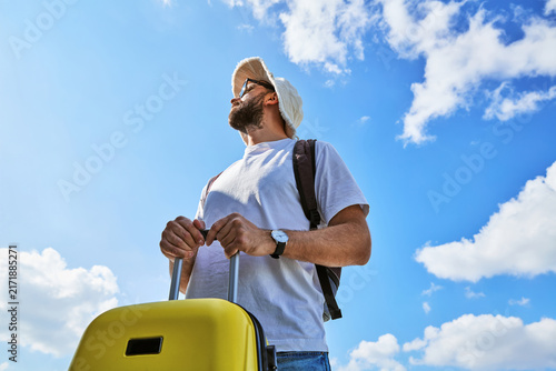 Canvastavla Male traveler with yellow suitcase and backpack looking up against blue sky