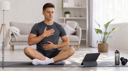 Young Man Practicing Mindful Deep Breathing Meditation on a Yoga Mat in a Bright Living Room, Following Online Wellness Tutorial
