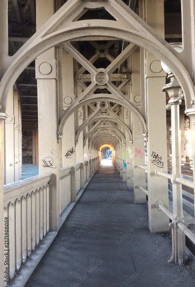 Fototapeta premium Long pedestrian walkway beneath a metal bridge with repeating structural arches forming a dramatic symmetrical perspective toward a distant opening. 