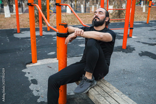 Fotografie Adult Man Sits Quietly On Orange Calisthenics Equipment In Urban Park, Eyes Clos