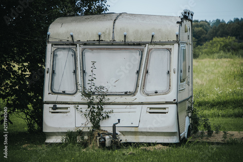 Фотография A white, weather-worn caravan standing under trees in a grassy rural field, with weeds growing around its exterior, a sign of abandonment and decay