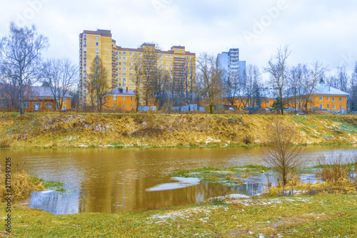 Fotografie Tall yellow apartment block and smaller yellow houses line riverbank with leafle