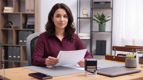 Brunette businesswoman notary holding document while sitting at wooden desk with stamp and laptop in office