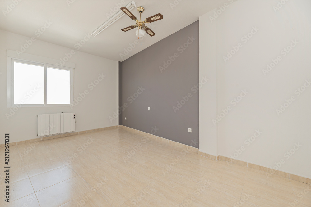 Fototapeta premium Empty room with gray accent wall, ceiling fan, window with radiator, and beige tiled floor