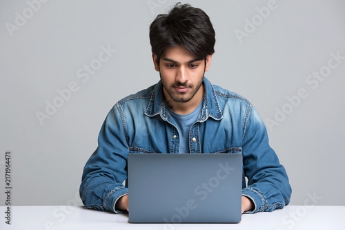 Young south asian man typing on a laptop computer against a plain grey studio background