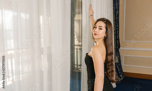 Bild auf Leinwand A beautiful young woman in an evening gown stands by a window with white curtains