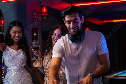 Quadro su tela A focused bearded DJ mixing on a controller while two women watch with glasses of wine presses a pad on the controller under the red neon strip at the DJ booth of a neon-lit nightclub