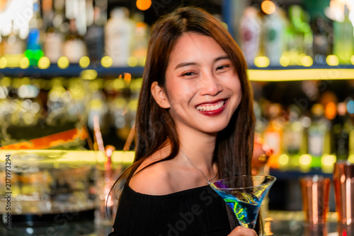 Фотография A beautiful young Asian woman in a black off-shoulder top holding a bright blue cocktail laughs with the cocktail glass tilted in her hand in a flattering beauty close-up
