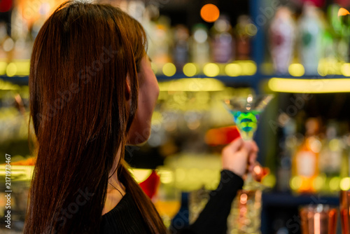 Cuadro en lienzo A beautiful young Asian woman in a black off-shoulder top holding a bright blue cocktail brings the blue martini slowly toward her lips at the polished counter of a cocktail bar