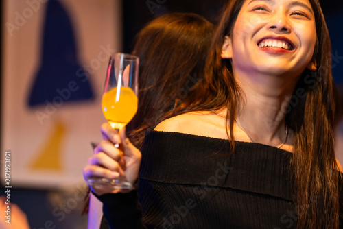 Quadro su tela Two stylish young Asian friends in black dresses talking over colourful cocktails smile with the drink lifted toward the camera in a stylish candlelit bar lounge