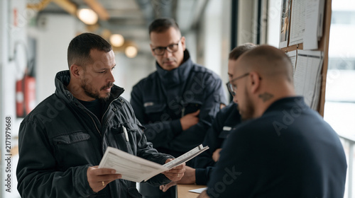 Freight forwarding agent cross references two shipping manifests in a hospital loading bay hallway, group of drivers gathered around a route board for a logistics road trip, quiet