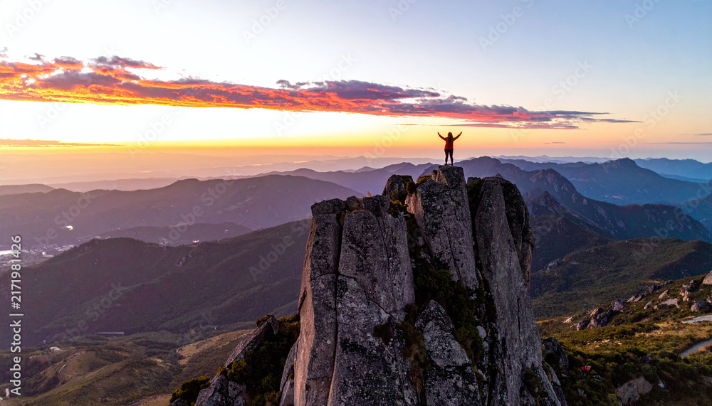 Naklejka premium A triumphant hiker stands atop a dramatic mountain peak with arms raised at vibrant sunset. An inspiring aerial view evoking freedom, achievement, and awe for travel concepts.