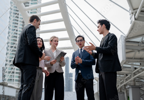 Diverse group of five business colleagues discussing project on urban skywalk, professional team meeting outdoors with tablet, city skyscrapers in background, teamwork concept